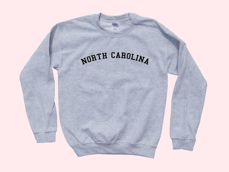 May include: A light gray sweatshirt with the text "NORTH CAROLINA" arched across the chest in black letters. The long-sleeved crew neck top is made of a soft material. The sweatshirt is a casual style.