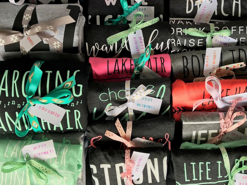 May include: A stack of folded t-shirts with various designs and colors, including black, gray, pink, green, and red. Each shirt is tied with a ribbon and a small tag with a message. The tags say "Happy Birthday", "Just Be Cause", "Congrats", and "Lake Hair".