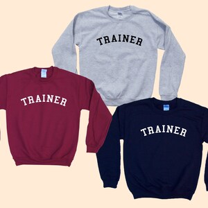 Pu&ograve; includere: Tre felpe girocollo bordeaux, grigie e blu navy. Ogni felpa ha la scritta "TRAINER" stampata in bianco, con lettere arcuate sul petto. Le felpe sono esposte su uno sfondo beige chiaro.