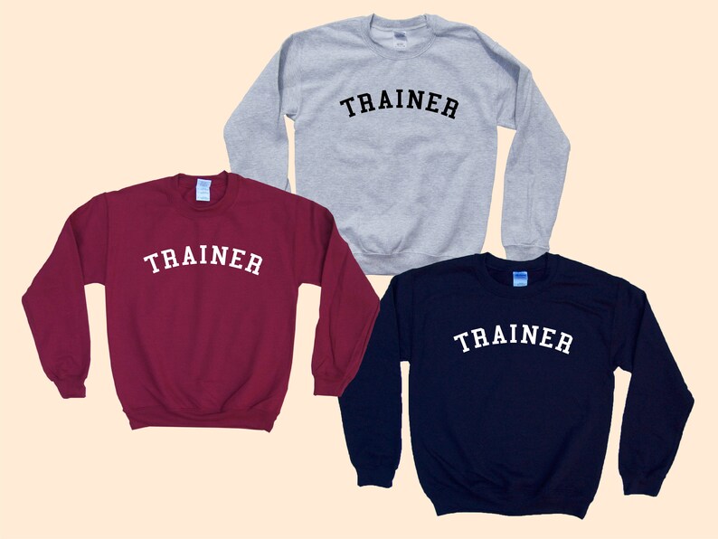 Pu&ograve; includere: Tre felpe girocollo bordeaux, grigie e blu navy. Ogni felpa ha la scritta "TRAINER" stampata in bianco, con lettere arcuate sul petto. Le felpe sono esposte su uno sfondo beige chiaro.