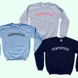 May include: Three sweatshirts in different colours: light blue, grey, and navy blue. Each sweatshirt has the word "STATISTICS" printed on the front in a black arched font.