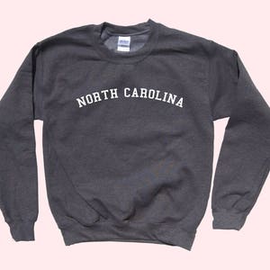 May include: A dark gray sweatshirt with the text "NORTH CAROLINA" in white, arched across the chest. The long-sleeved crewneck top is made of a soft material and is suitable for casual wear.