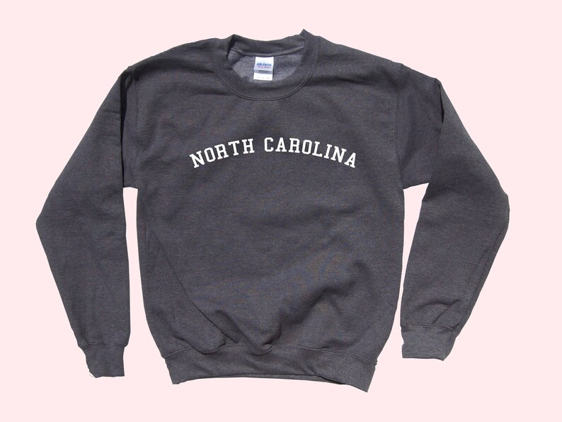 May include: A dark gray sweatshirt with the text "NORTH CAROLINA" in white, arched across the chest. The long-sleeved crewneck top is made of a soft material and is suitable for casual wear.