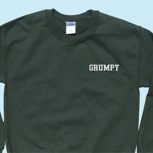 Peut inclure: Sweat-shirt vert foncé avec le mot "GRUMPY" imprimé en blanc. Le sweat-shirt a un col rond et des manches longues. Le fond est bleu clair.