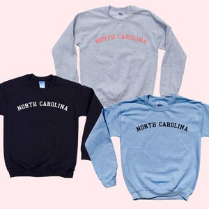 May include: Three sweatshirts in different colors: black, light blue, and gray. Each sweatshirt has the text "NORTH CAROLINA" printed across the chest in a contrasting color. The sweatshirts are displayed on a light pink background.
