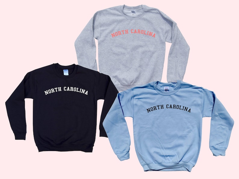May include: Three sweatshirts in different colors: black, light blue, and gray. Each sweatshirt has the text "NORTH CAROLINA" printed across the chest in a contrasting color. The sweatshirts are displayed on a light pink background.