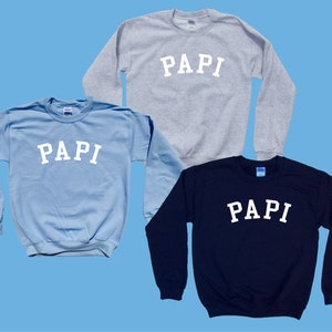 May include: Three crewneck sweatshirts in different colors: light blue, gray, and navy blue. Each sweatshirt has the word "PAPI" printed in white on the front.