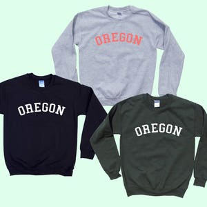 May include: Three sweatshirts in different colors, gray, black, and green, with the word "OREGON" printed in white on each. The word "OREGON" is in an arched font.