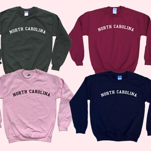 May include: Four crewneck sweatshirts in different colors: dark green, burgundy, light pink, and navy blue. Each sweatshirt has the text "NORTH CAROLINA" printed in white, arched across the chest. The sweatshirts are displayed on a light pink background.