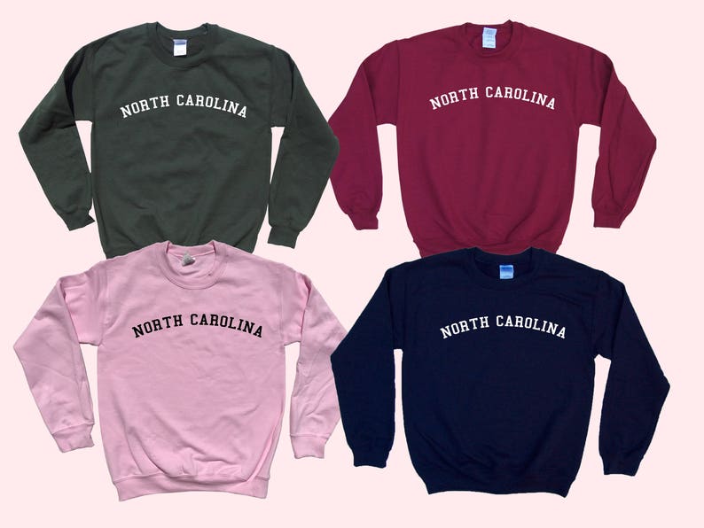 May include: Four crewneck sweatshirts in different colors: dark green, burgundy, light pink, and navy blue. Each sweatshirt has the text "NORTH CAROLINA" printed in white, arched across the chest. The sweatshirts are displayed on a light pink background.