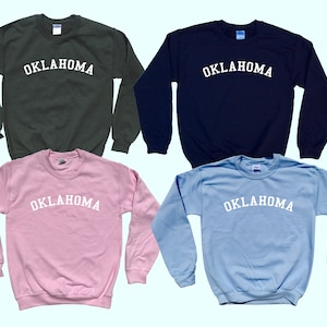 May include: Four sweatshirts in different colors: dark green, navy blue, light pink, and light blue. Each sweatshirt has the word "OKLAHOMA" printed in white on the front.