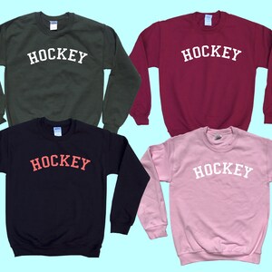 Pu&ograve; includere: Quattro felpe di diversi colori con la parola "HOCKEY" stampata in un arco bianco sul davanti. Le felpe sono di colore verde, bordeaux, nero e rosa.
