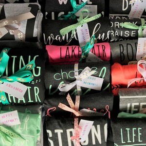 May include: A stack of folded t-shirts with various designs and colors, including black, gray, pink, green, and red. Each shirt is tied with a ribbon and a small tag with a message. The tags say "Happy Birthday", "Just Be Cause", "Congrats", and "Lake Hair".