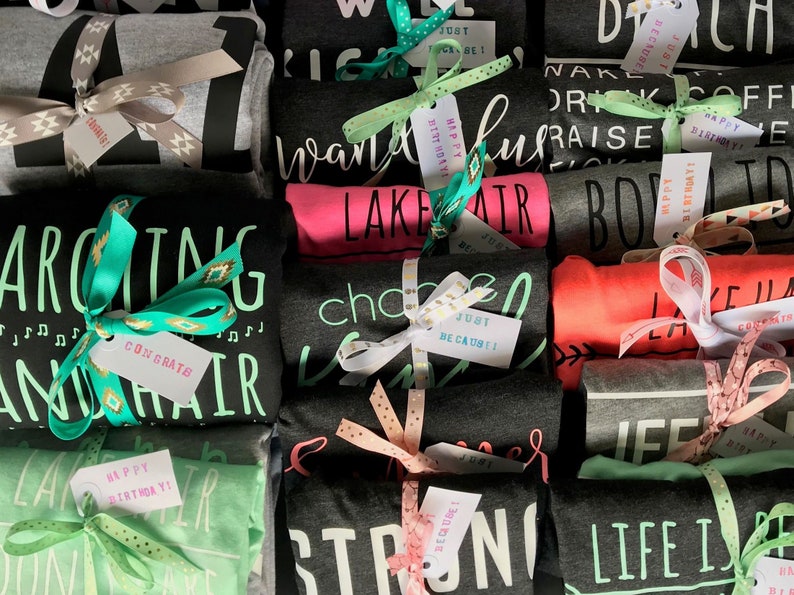 May include: A stack of folded t-shirts with various designs and colors, including black, gray, pink, green, and red. Each shirt is tied with a ribbon and a small tag with a message. The tags say "Happy Birthday", "Just Be Cause", "Congrats", and "Lake Hair".