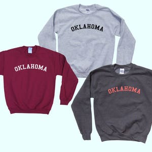 May include: Three sweatshirts in different colors with the word "OKLAHOMA" printed in a black, white, and red arch across the front.