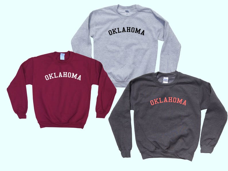 May include: Three sweatshirts in different colors with the word "OKLAHOMA" printed in a black, white, and red arch across the front.