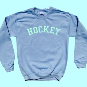 Pu&ograve; includere: Una felpa blu chiaro con la parola "HOCKEY" stampata in verde chiaro sul petto.