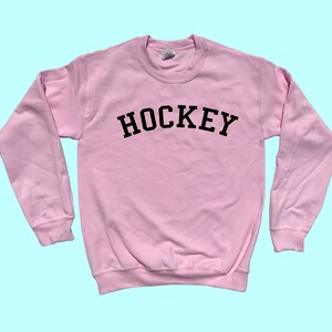 Pu&ograve; includere: Una felpa rosa con la scritta "HOCKEY" stampata in nero sul davanti.