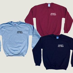 May include: Three sweatshirts in different colors: light blue, maroon, and navy blue. Each sweatshirt has the text "GRUMPY OLD MAN" embroidered on the front in white.
