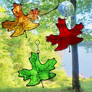 May include: Three stained glass maple leaves, one red, one green, and one yellow-orange, are hanging on a window screen.