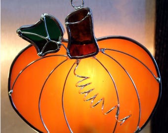 May include: A stained glass pumpkin nightlight featuring a bright orange body with silver metal soldering lines, a dark green leaf, and a brown stem. A silver metal spiral wire accent is attached to the front, and a white plug base is visible.