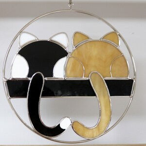 May include: A stained glass suncatcher featuring two cats, one black and white and the other orange, with their tails intertwined. The cats are positioned as if they are sleeping, with their backs to the viewer. The suncatcher is circular and has a silver frame.