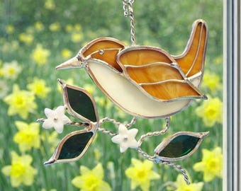 May include: Stained glass bird suncatcher with a brown and cream body, perched on a silver wire branch with green leaves and white flowers. The bird is hanging from a suction cup.