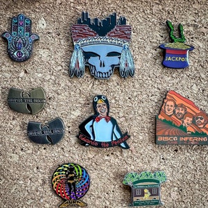 May include: A collection of enamel pins featuring various designs, including a skull with a headdress, a penguin, a hand with an eye, a mushroom, a building, and a mountain range. The pins are all different colors and sizes.