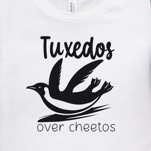 Könnte beinhalten: Weißes T-Shirt mit schwarzem Text, der "Tuxedos over Cheetos" lautet, und einer schwarzen Silhouette eines Pinguins, der einen Smoking trägt und eine Tüte Cheetos hält.