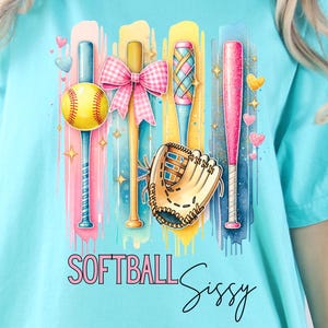 May include: A turquoise blue t-shirt with a graphic design of colorful paint splatters, baseball bats, a softball, and a glove. The text "SOFTBALL Sissy" is printed below the graphic.