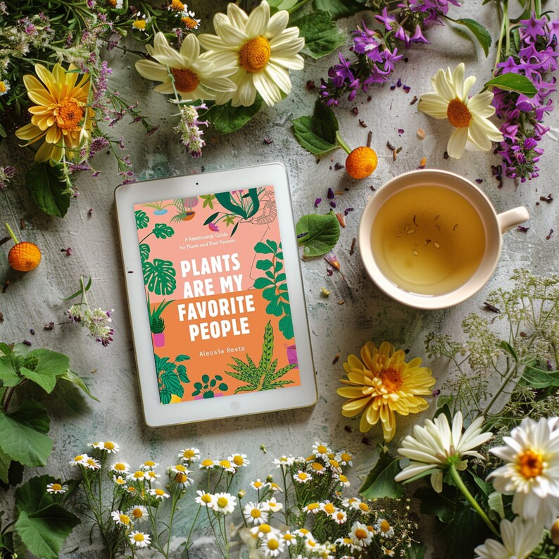 May include: A book titled "Plants Are My Favorite People: A Handbook to Grow Plants and Grow Your Own Happiness" by Alexia Festa. The book cover features a colorful illustration of various plants and flowers. The book is lying on a table with a cup of tea and flowers.