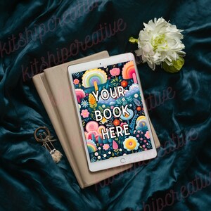 K&ouml;nnte beinhalten: Ein wei&szlig;es Tablet mit einem bunten Blumenmuster und dem Text "YOUR BOOK HERE" auf dem Bildschirm. Das Tablet liegt auf einem Stapel beiger B&uuml;cher auf einer dunkelblauen Oberfl&auml;che.