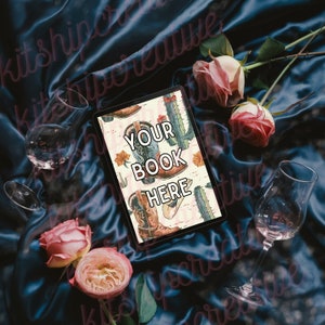 K&ouml;nnte beinhalten: Ein digitales Tablet mit einem rosa und wei&szlig;en Hintergrund, der ein Muster aus Cowboystiefeln und Kakteen zeigt. Der Text "YOUR BOOK HERE" wird auf dem Bildschirm angezeigt. Das Tablet liegt auf einem blauen Satin-Stoff mit rosa Rosen und Weingl&auml;sern.