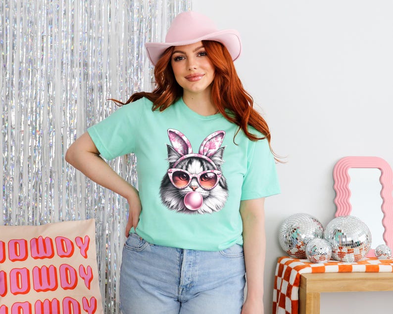 K&ouml;nnte beinhalten: Eine Frau tr&auml;gt ein hellgr&uuml;nes T-Shirt mit einem Aufdruck eines Katers mit Hasenohren und Sonnenbrille, der eine Blase bl&auml;st. Der Kater tr&auml;gt rosa Hasenohren und rosa Sonnenbrille. Die Frau tr&auml;gt auch einen rosa Hut und hat lange rote Haare.