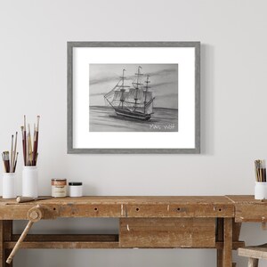 Puede incluir: Dibujo a l&aacute;piz enmarcado de un velero navegando en el mar, firmado "Marc Wolf". La obra de arte est&aacute; en un marco gris. La imagen se muestra en un banco de trabajo de madera con materiales de arte.