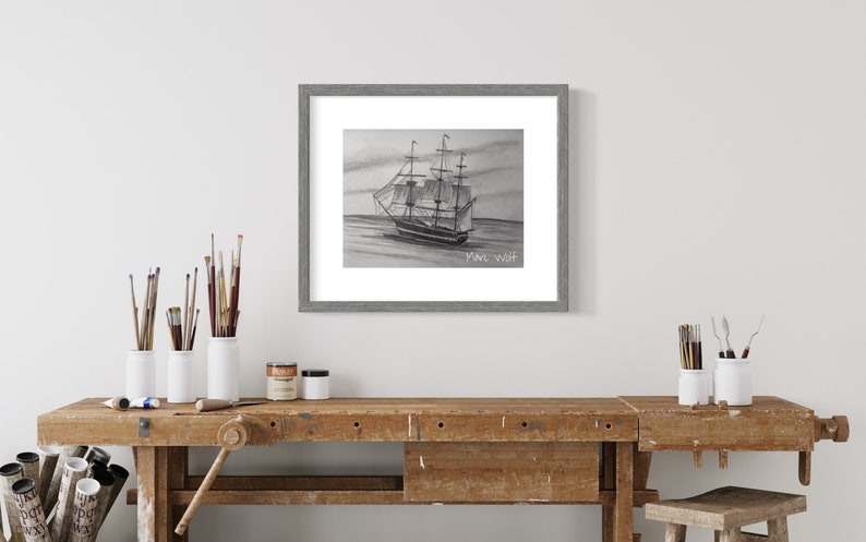 Puede incluir: Dibujo a l&aacute;piz enmarcado de un velero navegando en el mar, firmado "Marc Wolf". La obra de arte est&aacute; en un marco gris. La imagen se muestra en un banco de trabajo de madera con materiales de arte.