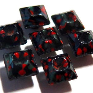 May include: Seven square beads with faceted edges. The beads are dark teal with red speckles and a hole in the center. The beads are arranged on a white surface, casting shadows.