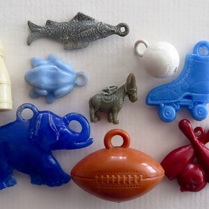 May include: A collection of nine plastic charms, including a blue frog, a blue elephant, a brown football, a red and white baseball, a blue roller skate, a brown donkey, a red and white bowling pin set, a grey fish, and a white bottle.
