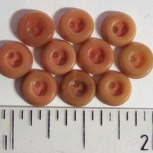 Könnte beinhalten: Zehn braune Plastikknöpfe mit jeweils zwei Löchern. Die Knöpfe haben eine leicht erhabene Form in der Mitte.