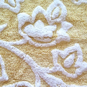 May include: Close-up of a yellow bath mat with a floral design in white. The mat has a textured, plush surface. The white floral pattern is raised, creating a three-dimensional effect. The mat is likely made of absorbent material.