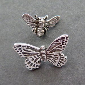 Può includere: Due spille in metallo color argento a forma di insetti. La spilla superiore è un'ape con ali dettagliate e un corpo segmentato. La spilla inferiore è una farfalla con intricati motivi sulle ali e un corpo centrale ben definito.