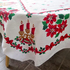 May include: White Christmas tablecloth with red poinsettias, green leaves, and red bows. The tablecloth features a design of three red candles and gold candle holders. The edges are decorated with a ribbon of red bows and holly.