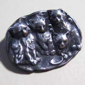 May include: An oval-shaped silver brooch featuring a relief design of three cats sitting together. The metallic surface has a polished finish with darkened recessed areas that highlight the detailed fur textures and facial features of the cats.