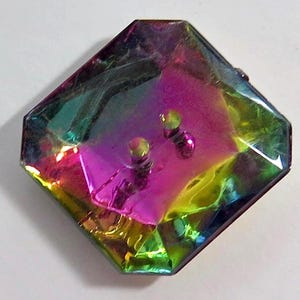 Op de afbeelding: Iridescent, veelkleurige knoop met een ruitvorm. De knoop heeft een levendige weergave van kleuren, waaronder roze, groen en geel. Twee kleine, zilverkleurige pinnen zijn zichtbaar op het oppervlak van de knoop.