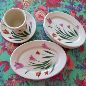 May include: A set of white ceramic dishes with a floral design. The set includes a teacup and saucer, plus several oval-shaped plates. The floral design features pink and orange tulips with green stems and leaves.