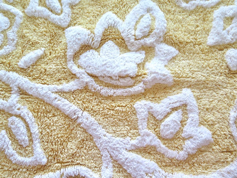May include: Close-up of a yellow bath mat with a floral design in white. The mat has a textured, plush surface. The white floral pattern is raised, creating a three-dimensional effect. The overall design is soft and inviting.