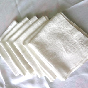 Può includere: Un set di sei tovaglioli di lino bianco sporco. Ogni tovagliolo quadrato è ben piegato e presenta un bordo cucito. I tovaglioli sono disposti a ventaglio su una superficie bianca, mostrando il loro design pulito e semplice.