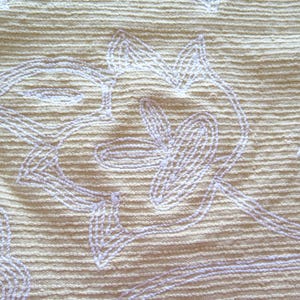 May include: Close-up of a textured, beige fabric with a repeating floral pattern embroidered in a light lavender color. The embroidery features stylized leaves and curved lines, creating a delicate, handcrafted appearance.