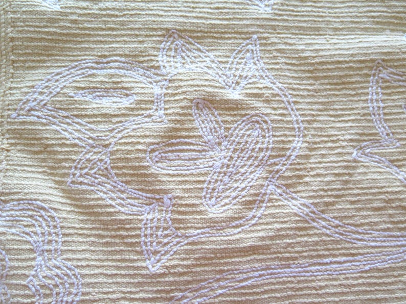 May include: Close-up of a textured, beige fabric with a repeating floral pattern embroidered in a light lavender color. The embroidery features stylized leaves and curved lines, creating a delicate, handcrafted appearance.
