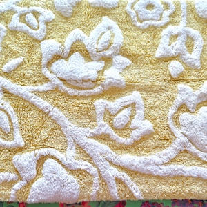 May include: Close-up of a yellow bath mat with a white floral design. The mat has a textured, plush surface with raised white floral patterns. The design features stylized flowers and flowing lines.